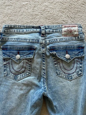 True Religion ljusblå jeans - Säljer ett par ljusblå jeans från True Religion med ikoniska bakfickor, röda kontrastsömmar och silverfärgade knappar. Jeansen har en klassisk femficksmodell och patch med logga bak i midjan. Perfekta för en cool och avslappnad stil.
