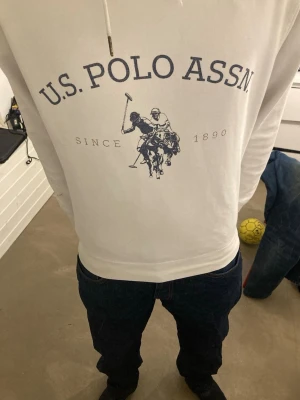 Vit hoodie från U.S. Polo Assn. - Vit hoodie från U.S. Polo Assn. med blå logga och tryck framtill. Klassisk huva med dragsko och långärmad modell. Perfekt för en avslappnad och sportig stil.