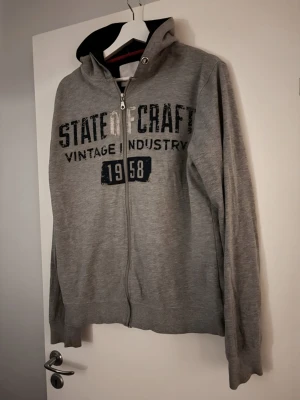 Grå hoodie med tryck och dragkedja - Snygg grå hoodie från lager 157med svart tryck på bröstet där det står 'Vintage Industry 1958'. Hoodien har dragkedja framtill, huva med svart insida och långa ärmar. Perfekt för en avslappnad och cool stil.