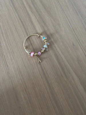 Guldigt armband med färgglada pärlor - Snyggt guldigt armband med flera färgglada pärlor i rosa, lila och blått. Armbandet har även berlocker, bland annat en liten Eiffeltorn-berlock och glittrande detaljer. Perfekt accessoar för dig som gillar unika smycken med charm.