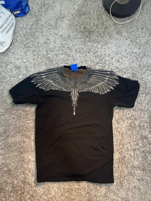 Svart Marcelo Burlon t-shirt  - Snygg svart t-shirt från Marcelo Burlon med ikoniskt vingtryck i grått och vitt runt halsen. T-shirten har en klassisk passform och är kortärmad, perfekt för en cool och edgy stil.