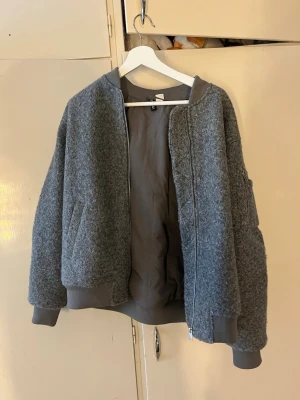 Grå bomberjacka  - Snygg grå bomberjacka med lång ärm och ribbade muddar. Jackan har dragkedja framtill och en extra ficka med dragkedja på ärmen. Tillverkad i ullblandning för en varm och trendig look.