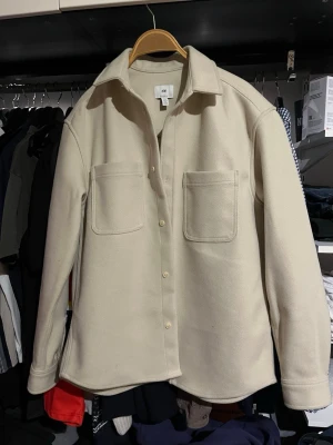 Beige overshirt från H&M - Stilren beige overshirt från H&M med klassisk krage och två stora bröstfickor. Skjortjackan har raka ärmar, knäppning framtill och är tillverkad i ett mjukt, filtliknande material. Perfekt för lager-på-lager och enkel att matcha med olika outfits.
