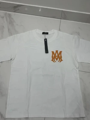 Vit AMIRI t-shirt med orange tryck - Snygg vit t-shirt från AMIRI med orange logga på bröstet och stort AMIRI-tryck på ryggen. Klassisk rund hals och korta ärmar. Tillverkad i mjuk bomull som känns skön mot huden. Perfekt för dig som gillar streetwear och vill sticka ut.