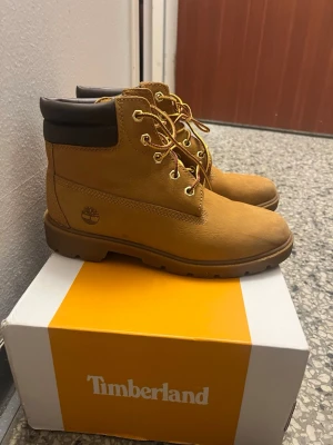 Timberland klassiska gula kängor - Timberland kängor i klassisk gul nubuck med bruna detaljer vid ankeln och robust gummisula. Kängorna har metallöglor, gulbruna snören och ikoniska Timberland-loggan på sidan. Perfekta för höst och vinter med sin slitstarka design.