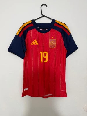 Spaniens matchtröja Lamine Yamal - Röd och marinblå Spanien matchtröja från Adidas med gula detaljer och nummer 19. Tröjan har Lamine Yamal tryckt på ryggen, korta ärmar och klassiska Adidas-ränder på axlarna. Perfekt för fotbollsfans som vill stötta landslaget.