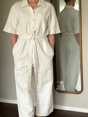 Vit kortärmad jumpsuit med knytskärp från COS - Stilren vit jumpsuit med korta ärmar, knappar framtill och  knytskärp i midjan. Modellen har en bröstficka och vida ben som slutar vid ankeln. Tillverkad i ett mjukt, vävt bomullstyg som känns luftigt och bekvämt. Från COS. 