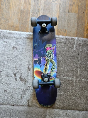 Skateboard med färggrafik - Cool skateboard med färgglad grafik på undersidan som visar en robotliknande figur med rosa hårband. Brädan har svarta hjul och svart griptape på ovansidan. Perfekt för dig som gillar unika motiv och vill sticka ut i skateparken. Använt skik, tecken på slitage