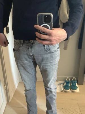 Grå jeans från Replay 31/32 modell anbass - Snygga grå jeans från Replay med klassisk femficksdesign och tydliga svarta sömmar på bakfickorna. Jeansen har en ljusgrå tvätt och ett stort Replay-märke på linningen. Perfekta för en avslappnad och trendig stil.