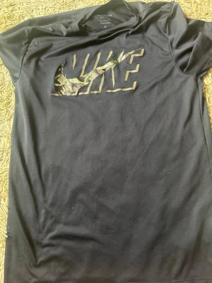 Svart Nike Dri-Fit t-shirt med camologga - Svart t-shirt från Nike med Dri-Fit-material och stor logga framtill i camouflage-mönster. Klassisk rund halsringning och korta ärmar. Perfekt för dig som gillar sportig och stilren look. Har nog aldrig använt den.