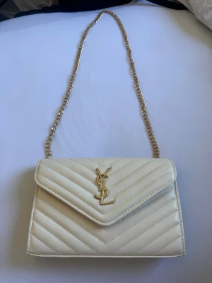 Vit axelväska  - Snygg axelväska i krämvitt läder från Saint Laurent med guldfärgad kedja och YSL-logga framtill. Köpt från Turkiet. Pris kan diskuteras.
