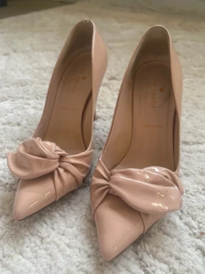 Klackskor Kate Spade - Snygga nude-färgade pumps från Kate Spade i glansigt lackmaterial. Skorna har spetsig tå och en dekorativ rosett framtill. Klackarna är smala och höga, vilket ger en elegant siluett. Perfekta för dig som gillar stilrena och trendiga detaljer.