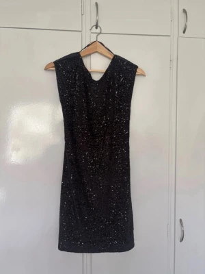 Svart glittrig kortklänning - Snygg svart kortklänning med glittriga paljetter och öppen rygg. Klänningen är ärmlös och har en figurnära passform som ger en elegant look. Perfekt för dig som vill sticka ut på nästa fest! Materialet känns mjukt och följsamt.