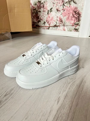 Nike Air Force 1 vita sneakers - Klassiska vita Nike Air Force 1 sneakers i lågt utförande. Skorna har en stilren design med perforerad tå, vit sula och Nike-logga på sidan och hälen. Tillverkade i slätt läder med vita skosnören och metallbricka på snörningen. Perfekta för en clean och tidlös look.