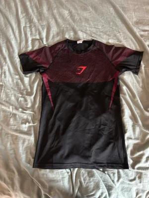 Svart och vinröd träningströja Gymshark - Snygg svart och vinröd träningströja från Gymshark med korta ärmar. Tröjan har ett sportigt mönster i vinrött över bröst och axlar samt Gymshark-logga framtill. Tillverkad i ett lätt och stretchigt funktionsmaterial som passar perfekt till gymmet.