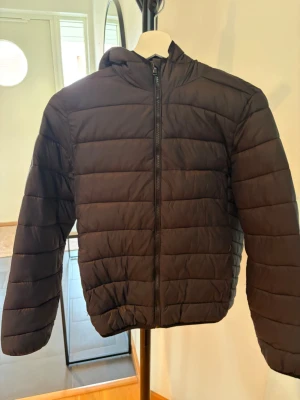 Svart pufferjacka med huva - Snygg svart pufferjacka med huva och dragkedja framtill. Jackan har quiltad design med horisontella sömmar och elastiska muddar vid ärmarna. Perfekt för kyliga dagar och enkel att matcha till olika outfits.