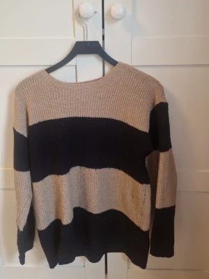 Beige och svart randig stickad tröja - Stickad tröja från 157 med breda horisontella ränder i beige och svart. Tröjan har rund halsringning och långa ärmar. Perfekt för dig som gillar en avslappnad och stilren look.