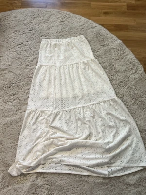 Vit långkjol med broderade detaljer - Superfin vit långkjol från Kappahl med broderade hål och volangiga partier. Kjolen har resår i midjan och är gjord i mjuk bomull, perfekt för en somrig look. Snygg att matcha med en enkel topp eller stickad tröja.