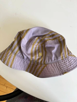 Lila bucket hat med guldiga ränder - Snygg bucket hat i lila med guldiga ränder och liten fyrkantig logga framtill. Hatten har klassisk form och är tillverkad i ett lätt tyg, perfekt för att lyfta din stil. Passar dig som gillar trendiga accessoarer och vill sticka ut.