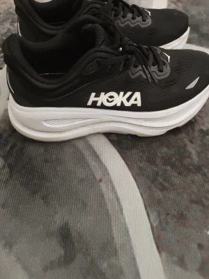 Svarta Hoka sneakers med vit sula - Svarta sneakers från Hoka med vit, tjock sula och svart ovandel i mesh. Skorna har vita detaljer och Hoka-logga på sidan. Klassisk snörning och låg modell som passar perfekt till en sportig stil.