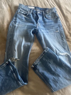 Ljusblå slitna raka jeans - Säljer ett par ljusblå jeans med raka ben och slitningar vid knäna och låret. Klassisk femficksmodell med hög midja och knappgylf. Jeansen har en avslappnad passform och är tillverkade i denim av bomull.
