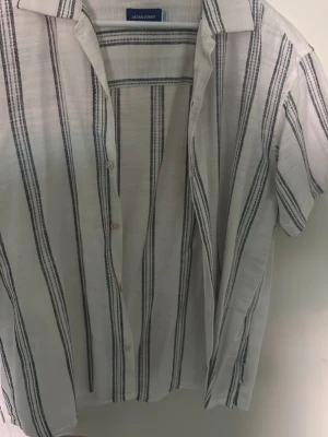 Randig skjorta från Jack & Jones använd 1 gång - Vit skjorta från Jack & Jones med svarta vertikala ränder. Skjortan har korta ärmar, klassisk krage och knäppning framtill. Tillverkad i ett lätt och luftigt material som passar perfekt till varmare dagar.