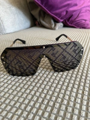 Fendi solglasögon med logomönster - Snygga solglasögon från Fendi med stort, sköldformat glas och ikoniskt Fendi-logomönster i guld och svart. Glaset är mörkt och har dekorativa nitar längs kanterna. Perfekt statement-piece för dig som vill sticka ut.