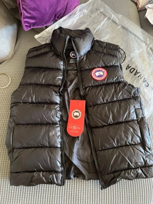 Svart dunväst från Canada Goose - Snygg svart dunväst från Canada Goose med klassisk logotyp på bröstet och hög krage. Västen har quiltad design, dragkedja framtill och är fylld med dun för extra värme. Perfekt för lager-på-lager och streetstyle.