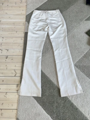 Vita utsvängda kostymbyxor  - Säljer ett par vita kostymbyxor med utsvängda ben och pressveck. Byxorna har fickor fram och bak samt hällor för skärp. Perfekta för en clean och stilren look. Materialet känns mjukt och följsamt.
