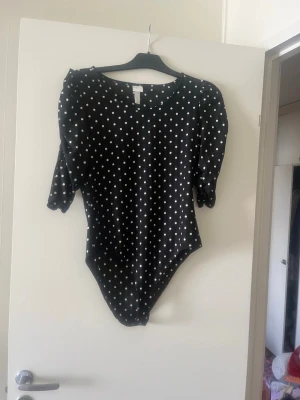 Svart prickig body med puffärm - Snygg svart body med vita prickar och puffärmar. Rund halsringning och trekvartsärm ger en trendig look. Tillverkad i mjukt stretchigt material som sitter skönt mot kroppen. Perfekt för dig som gillar en lekfull och stilren stil.