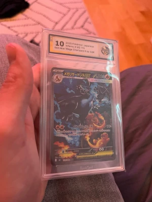 JapanskPokémon Mega Charizard X ex SAR kort - Japanskt samlarkort från 2025 med Mega Charizard X ex SAR-motiv. Kortet är graderat och inneslutet i ett genomskinligt plastskydd med etikett. Färgerna är svart, blå, orange och gult med detaljerad grafik och japansk text. Perfekt för Pokémon-entusiaster och samlare.