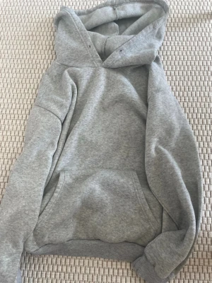 Grå hoodie med magficka - En klassisk grå hoodie med stor huva och magficka. Hoodien har ribbade muddar vid ärmslut och nederkant. Perfekt för en avslappnad stil och enkel att matcha med andra plagg. Materialet känns mjukt och bekvämt.