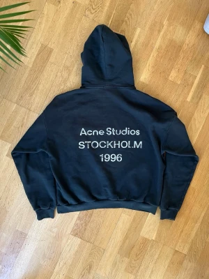 Svart hoodie Acne Studios Stockholm - Svart hoodie från Acne Studios med tryck på ryggen: 'Acne Studios STOCKHOLM 1996'. Klassisk passform med huva och ribbade muddar. Perfekt för en avslappnad och trendig streetwear-look. Storlek M men passar även L