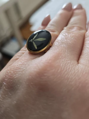 Svart oval ring med bladmotiv - Snygg oval ring med blad i guldton. Ringen har en guldfärgad ram vilket ger en elegant och stilren känsla. Perfekt accessoar för dig som gillar unika smycken med naturinspirerade detaljer. Material -rostfritt stål, justerbart.
