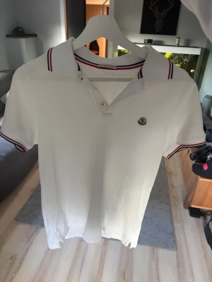 Vit Moncler pikétröja med rödblå detaljer - Snygg vit pikétröja från Moncler med klassisk krage och knäppning framtill. Tröjan har röda och blå ränder på krage och ärmslut samt Moncler-logga på bröstet. Perfekt för en stilren och sportig look. Nypris ligger runt 3500kr