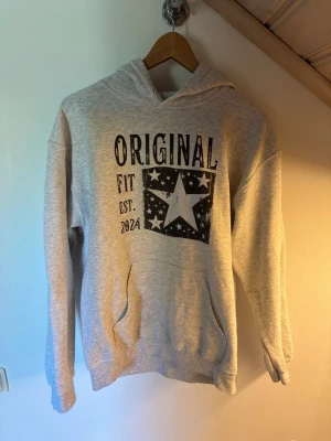 Grå hoodie med tryck och stjärna - Ljusgrå hoodie med svart tryck framtill där det står 'ORIGINAL FIT EST. 2024' och en stor stjärna omgiven av mindre stjärnor. Klassisk känguruficka och huva. Mjuk och skön bomullsblandning.