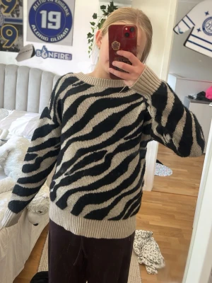 Beige och svart zebramönstrad stickad tröja - Stickad tröja med zebramönster i beige och svart. Tröjan har breda ribbade muddar vid ärmslut och nederkant samt en bred ribbad halsringning som kan bäras både som rundhalsad och v-ringad. Perfekt för dig som gillar djurmönster och oversized stil.