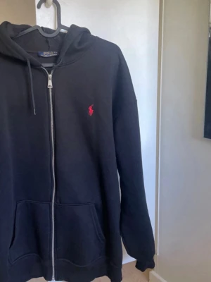 Svart hoodie från Polo Ralph Lauren - Svart hoodie med dragkedja från Polo Ralph Lauren. Klassisk design med röd broderad logga på bröstet, huva med dragsko och två stora fickor framtill. Tillverkad i mjukt material som är skönt att ha på sig. Perfekt för en avslappnad stil.
