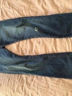 Blå Armani Jeans med slitningar - Säljer ett par blå jeans från Armani Jeans med snygga slitningar och en liten reva på låret. Klassisk femficksmodell med orangea sömmar och diskret logga på fickan. Jeansen har en rak passform och är tillverkade i denim.