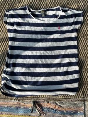 Randig t-shirt från Polo Ralph Lauren - Snygg t-shirt från Polo Ralph Lauren med breda marinblå och vita ränder. Klassisk passform med korta ärmar och rund hals. Liten rosa logga broderad på bröstet. Tillverkad i mjuk bomull, perfekt för en avslappnad stil.