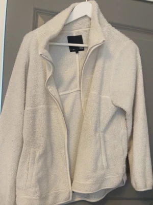 Vit teddyjacka  - Mysig vit teddyjacka från lager 157 med dragkedja framtill och två fickor. Jackan har hög krage och är gjord i fluffigt teddy-material som håller dig varm. Perfekt för kyliga dagar och enkel att matcha med olika outfits.