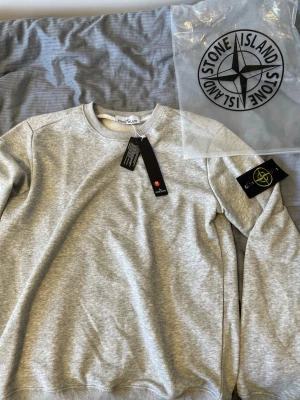 Grå sweatshirt från Stone Island - Snygg grå sweatshirt från Stone Island med klassisk rund hals och ribbade muddar. Tröjan har den ikoniska Stone Island-loggan på vänster ärm och kommer med originalpåse och tags kvar. Perfekt för en stilren och avslappnad look.