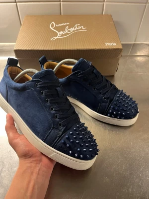Blåa Louboutin sneakers med nitar - Snygga blå sneakers från Christian Louboutin i mocka med coola matchande nitar på tån. Skorna har vita sulor, svarta skosnören och en klassisk låg modell. Perfekta för dig som vill sticka ut med en lyxig och edgy stil. Några spikar har gått av men medföljs extra spikar, som man kan lägga på om man vill