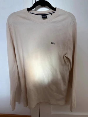 Beige långärmad tröja från BOSS - Stilren beige långärmad loungewear-tröja från BOSS Homewear. Tröjan har en diskret ribbad struktur och rund halsringning. Liten svart BOSS-logga broderad på bröstet. Perfekt för chill dagar hemma.