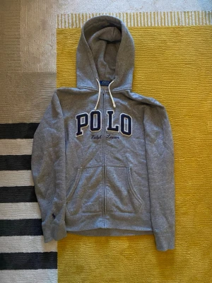 Grå hoodie från Polo Ralph Lauren - Snygg grå hoodie med dragkedja från Polo Ralph Lauren. Stor broderad POLO-text i blått och vitt framtill samt broderad logga under. Huva med vita snören och två fickor framtill. Perfekt för en avslappnad och sportig stil. Storlek S, ny pris 2300