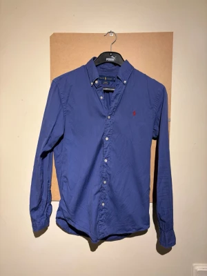Blå skjorta från Ralph Lauren - Klassisk blå skjorta från Ralph Lauren med button-down krage och lång ärm. Skjortan har vita knappar och den ikoniska röda logotypen broderad på bröstet. Tillverkad i bomull för en bekväm och stilren look.