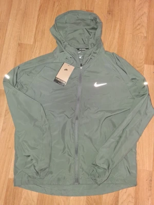 Grå Nike vindjacka med huva - Säljer en grå vindjacka från Nike med huva och hel dragkedja. Jackan har reflekterande detaljer och är tillverkad i lätt material. Loose fit och Move to Zero-kollektionen. Perfekt för att hålla vinden borta under utomhusaktiviteter.