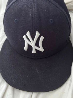 Mörkblå New York Yankees keps - Snygg mörkblå keps från New Era med klassiska New York Yankees-loggan broderad i vitt framtill. Kepsen har böjd skärm och ventilerande hål upptill. Perfekt accessoar för en sportig och urban stil. Skriv om ni är intresserade 