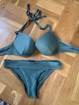 Grön ribbad bikini från H&M - Snygg grön bikini från H&M med ribbad struktur. Överdelen har vadderade kupor och knyt i nacken för en bekväm passform. Underdelen är klassisk med bred linning. Perfekt för stranddagar och poolhäng! Då jag köpte för stor storlek till mig så är bikinin aldrig använd. Storlek på bh:n är 85B och trosorna är 38. 