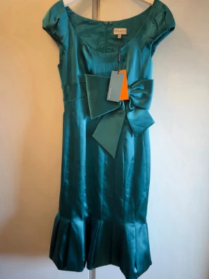 Turkos satin klänning med rosett - Supersnygg turkos festklänning från Karen Millen i glansigt satinmaterial. Helt ny med pislapp. Klänningen har korta ärmar, markerad midja och en stor rosett framtill. Kjolen har volangdetalj nertill för extra flärd. Perfekt för dig som vill sticka ut! Str 38 men liten mer som 36. 